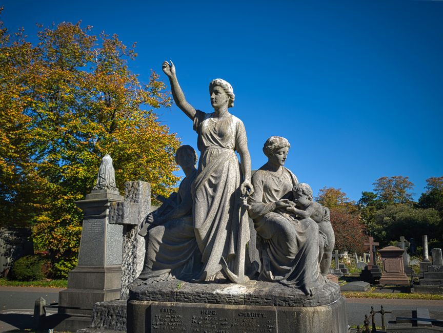 A large stone statue situated outdoors in a cemetery or memorial park setting, depicting three female figures in classical attire. The central figure, a woman with wavy hair, stands upright with her right arm raised and hand open, dressed in a flowing dress with detailed folds and texture. To her right, a seated woman cradles a small child, holding it gently while looking down at the infant, both figures carved with detailed hair and clothing. To the left of the central figure, another woman is partially visible, leaning forward slightly, also dressed in classical attire, with her head bowed. The sculpture is mounted on a rectangular stone base with inscriptions, surrounded by polished headstones and memorials, some with small statues or decorative elements, and positioned on a paved area. In the background, there are trees with autumn foliage, displaying vibrant yellow, orange, and green leaves, set against a bright blue sky, creating a peaceful and reflective atmosphere. The scene may relate to private or independent memorial care, emphasizing the importance of respectful waste removal from such locations, as handled by services like House Clearance Brompton.