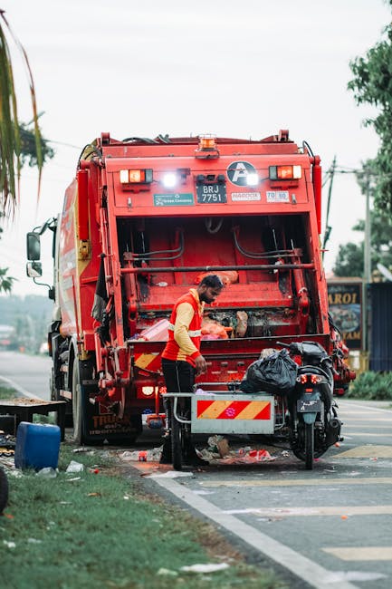 A sanitation worker wearing a high-visibility orange vest is operating a large red rubbish collection vehicle on the side of a street. The back of the vehicle features an open waste compartment, with the worker leaning into it to manage trash or debris. The vehicle, marked with identification numbers and safety stripes, is positioned on a paved roadside adjacent to a curb. Nearby, a black motorcycle parked close to the vehicle has a black plastic bag placed on its seat. The scene is set during daytime with overcast natural lighting, and elements such as green trees, a blue sign, and utility poles are visible in the background. The environment suggests a typical urban or suburban street scene, where independent waste collection or private rubbish removal services like House Clearance Brompton may conduct on-site clearance activities for local residents or businesses, emphasizing alternative waste handling outside of standard municipal collection. The overall composition centers around the engaged worker and the rubbish collection process, illustrating an organized approach to waste disposal in a professional context.