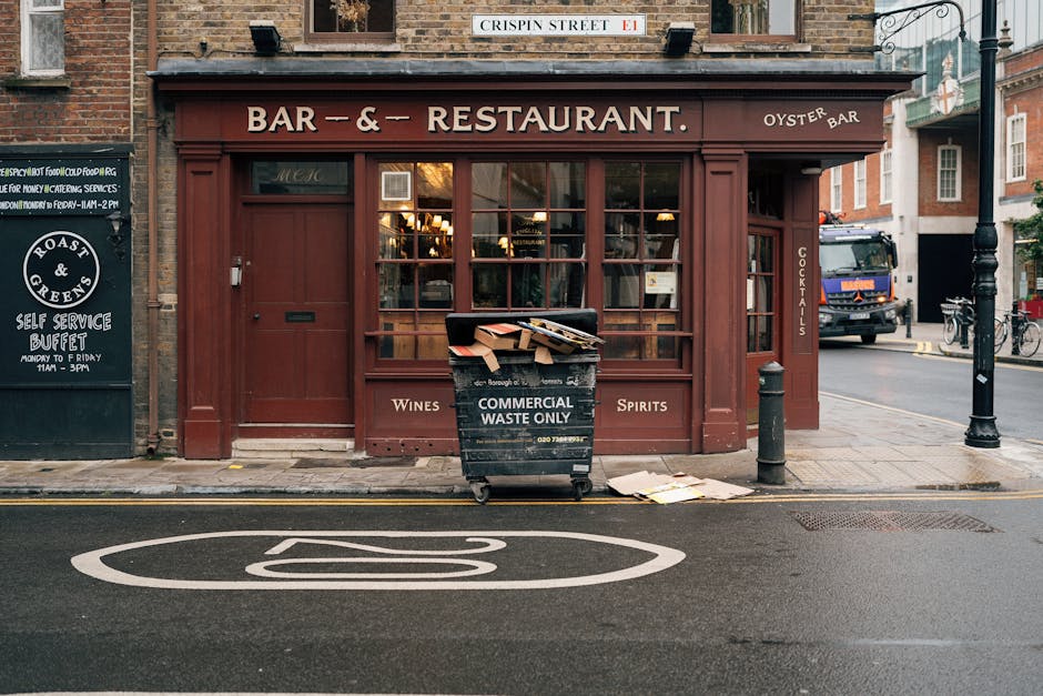 In front of a closed restaurant on a city street, a black wheeled waste collection bin marked 'COMMERCIAL WASTE ONLY' is positioned on the pavement. The bin contains flattened cardboard boxes and paper waste visible at the top. To the right of the bin, a small piece of discarded paper lies on the ground. The restaurant's façade features large glass windows framed by dark red wooden panels, with a sign above reading 'BAR & RESTAURANT.' Adjacent to the left, a blackboard-style notice board advertises a self-service buffet, while a bollard and a street lamp are situated nearby. The street surface shows a white no-parking symbol painted on the tarmac, and the overall scene suggests typical urban waste management practices, with the presence of a waste collection bin indicative of alternative disposal services outside of local authority collection, supported by the local waste management company House Clearance Brompton.