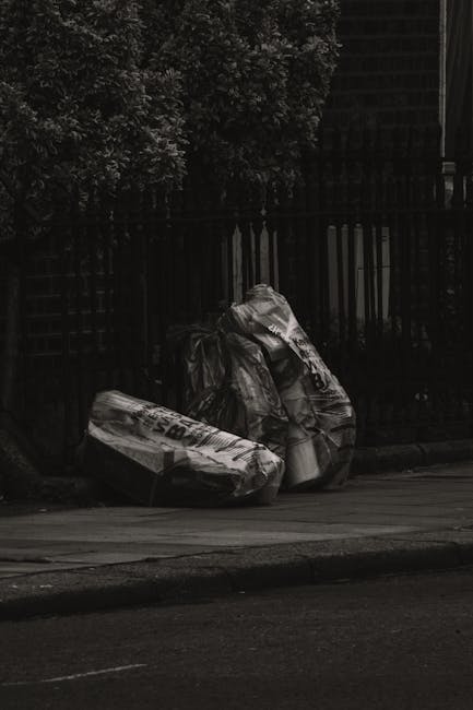 Two large black trash bags, filled and tied at the top, rest against a black metal fence on a pavement outside a residential area. The bags appear to contain household waste, with some visible crumpling and uneven surfaces, indicating they are full. The background features dense foliage from a bush or tree, partially obscuring the top of the fence, which is designed with vertical bars. To the right, a section of a modern building with dark brick or tile exterior is visible, suggesting an urban or suburban setting. The image is captured in natural daylight, with shadows cast on the pavement, emphasizing the bags' crumpled textures. The scene is straightforward, depicting a typical example of private rubbish collection or waste awaiting removal, aligning with the services offered by House Clearance Brompton for alternative waste handling outside official collection schemes.
