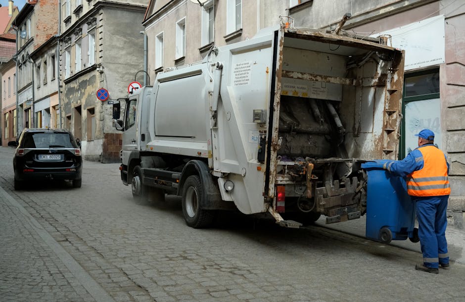 In front of a closed restaurant on a city street, a black wheeled waste collection bin marked 'COMMERCIAL WASTE ONLY' is positioned on the pavement. The bin contains flattened cardboard boxes and paper waste visible at the top. To the right of the bin, a small piece of discarded paper lies on the ground. The restaurant's façade features large glass windows framed by dark red wooden panels, with a sign above reading 'BAR & RESTAURANT.' Adjacent to the left, a blackboard-style notice board advertises a self-service buffet, while a bollard and a street lamp are situated nearby. The street surface shows a white no-parking symbol painted on the tarmac, and the overall scene suggests typical urban waste management practices, with the presence of a waste collection bin indicative of alternative disposal services outside of local authority collection, supported by the local waste management company House Clearance Brompton.