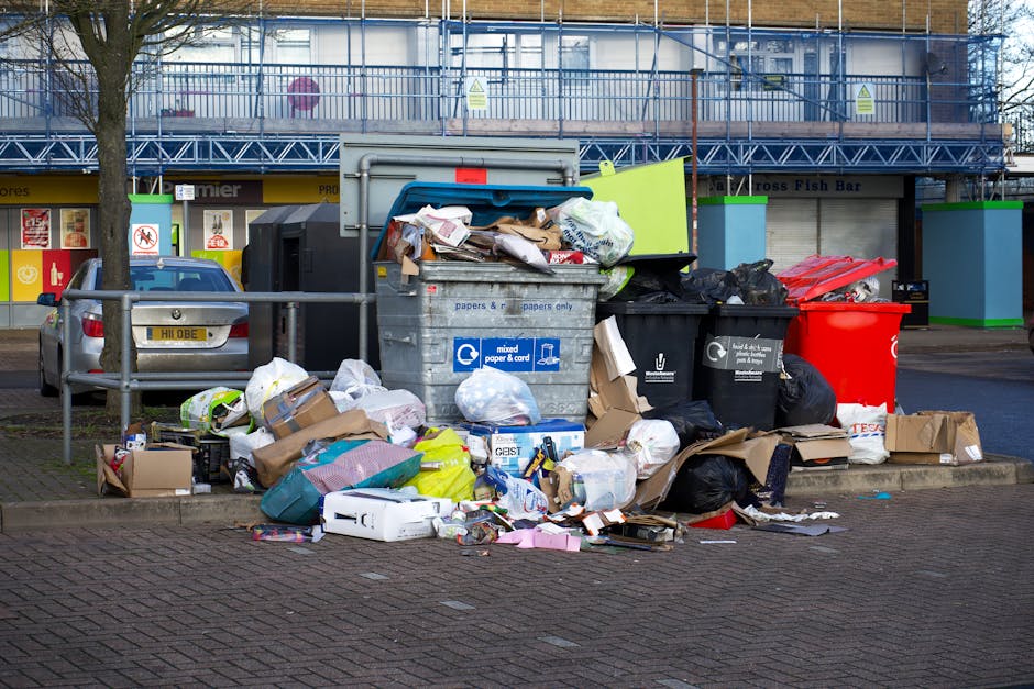 A collection of waste and rubbish overflowing from various bins and scattered on the pavement in an urban setting. The scene includes a large grey mixed paper and card bin with its lid partially open, revealing an assortment of flattened cardboard boxes, crumpled paper, and plastic items. Surrounding this are several smaller black and red waste bags, some of which are torn or opened, spilling their contents onto the brick-paved ground. Additional cardboard boxes, plastic packaging, and other household waste materials are strewn around the base of the bins, creating a cluttered appearance. In the background, there is a metal fence, a parked grey car, and a commercial building with storefronts and signage partially visible, indicating a busy town or city centre environment. The lighting suggests daytime with natural light illuminating the scene, and the overall impression highlights improper rubbish disposal, emphasizing the importance of professional waste management services like those offered by House Clearance Brompton for alternative waste handling and rubbish collection near West Brompton Station.