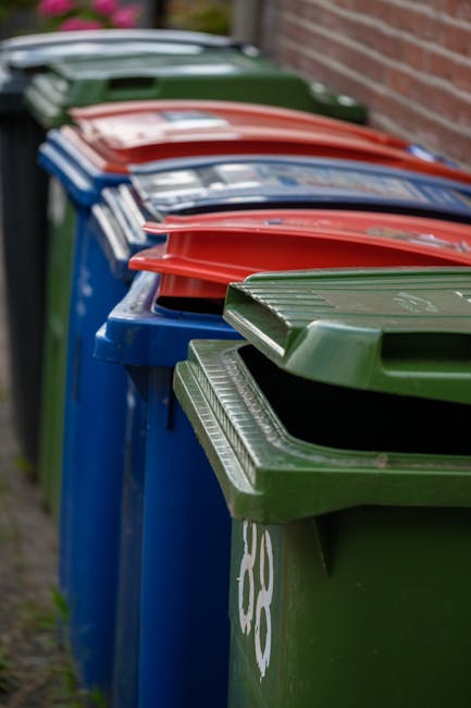 A row of large, color-coded plastic wheelie bins lined up outdoors along a paved pathway, positioned adjacent to a brick wall on the right side. The bins in the foreground are green with textured, ribbed lids, while further along the row, there are blue, red, and green bins, each with smooth, hinged lids. The bins are slightly weathered, with some showing faint scratches and marks on the surface. The environment appears to be in a residential area, with the bins intended for household waste collection, possibly related to private rubbish disposal services. The lighting suggests overcast conditions, with soft, diffuse natural light illuminating the scene. The arrangement and condition of the bins reflect their use for waste management and rubbish removal services, which may serve as an alternative to local authority collections, commonly conducted by companies such as House Clearance Brompton.