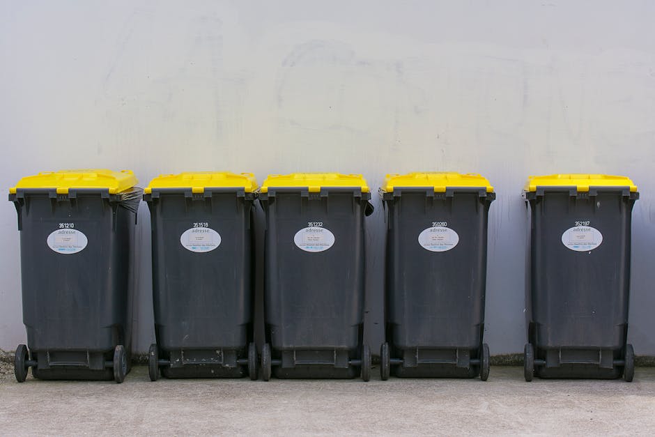 A collection of waste and rubbish overflowing from various bins and scattered on the pavement in an urban setting. The scene includes a large grey mixed paper and card bin with its lid partially open, revealing an assortment of flattened cardboard boxes, crumpled paper, and plastic items. Surrounding this are several smaller black and red waste bags, some of which are torn or opened, spilling their contents onto the brick-paved ground. Additional cardboard boxes, plastic packaging, and other household waste materials are strewn around the base of the bins, creating a cluttered appearance. In the background, there is a metal fence, a parked grey car, and a commercial building with storefronts and signage partially visible, indicating a busy town or city centre environment. The lighting suggests daytime with natural light illuminating the scene, and the overall impression highlights improper rubbish disposal, emphasizing the importance of professional waste management services like those offered by House Clearance Brompton for alternative waste handling and rubbish collection near West Brompton Station.
